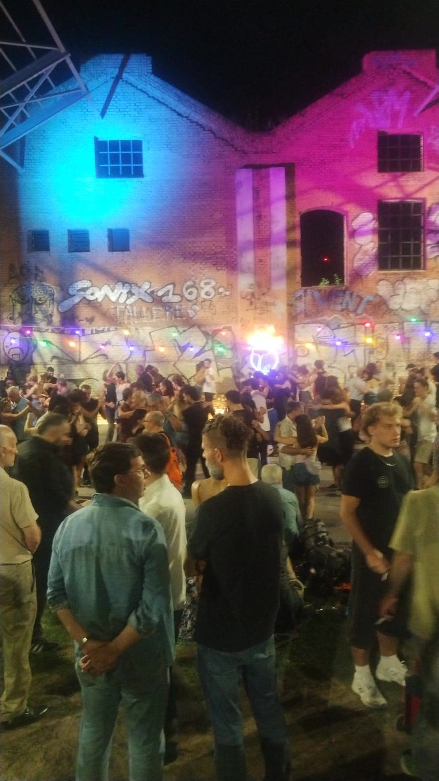 An open-air milonga at night, the crowd dancing in front of a graffiti-covered brick wall